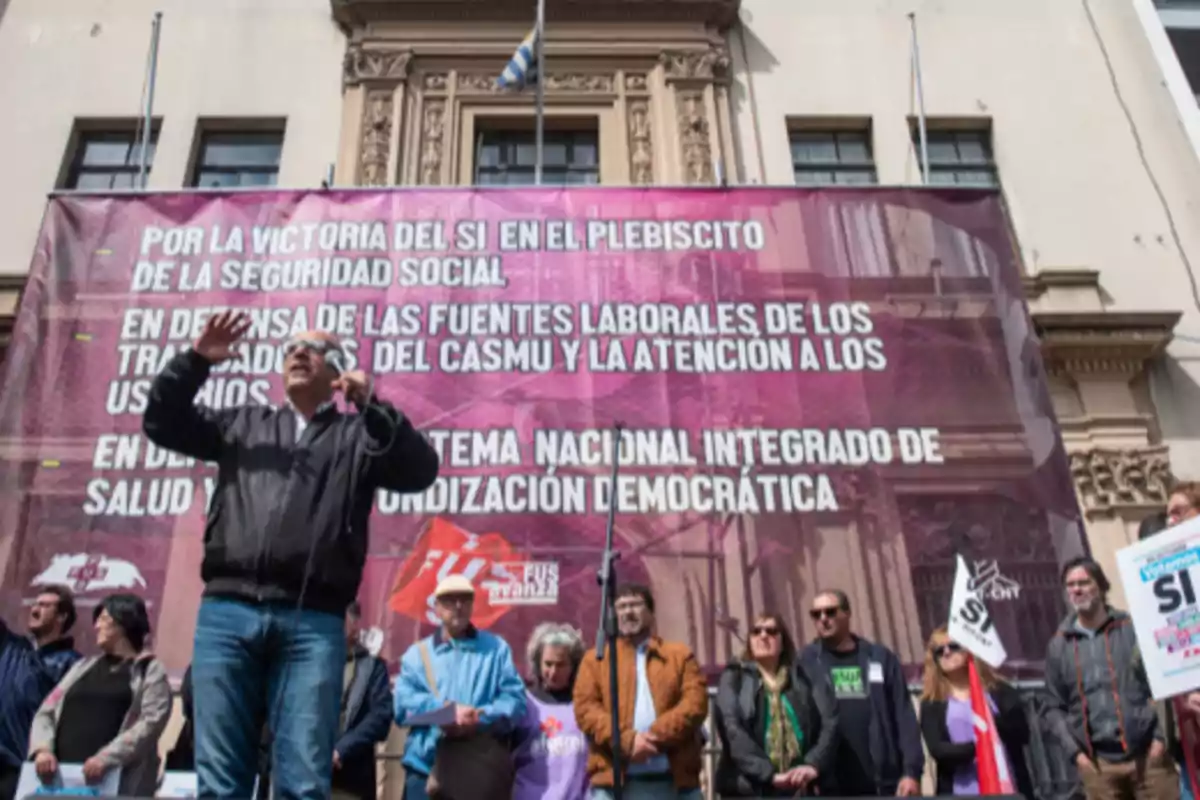 Un grupo de personas participa en una manifestación frente a un edificio, con un gran cartel de fondo que contiene mensajes en defensa de la seguridad social y el sistema de salud, mientras una persona habla por micrófono. Un grupo de personas participa en una manifestación frente a un edificio, con un gran cartel de fondo que contiene mensajes en defensa de la seguridad social y el sistema de salud, mientras una persona habla por micrófono.