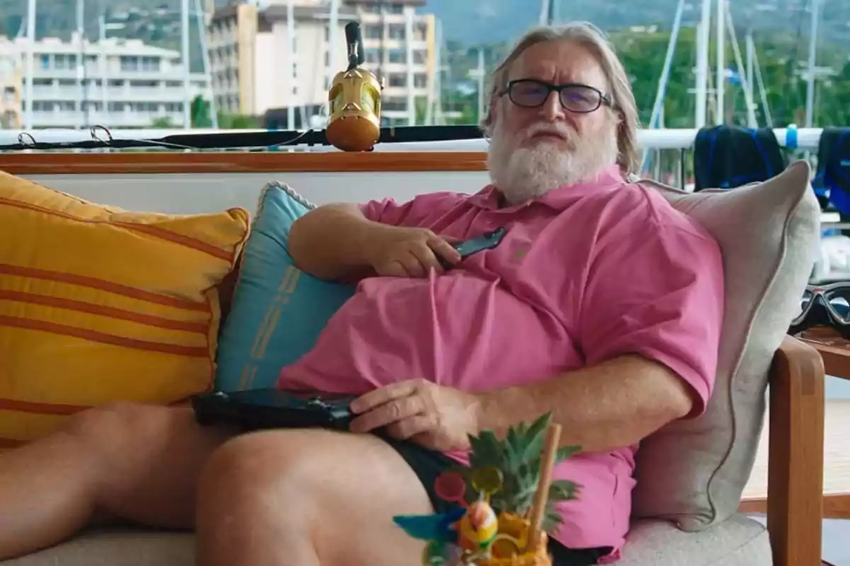 Hombre de barba y gafas sentado en un sofá al aire libre con una camisa rosa y un control remoto en la mano, rodeado de cojines y una bebida decorada en primer plano