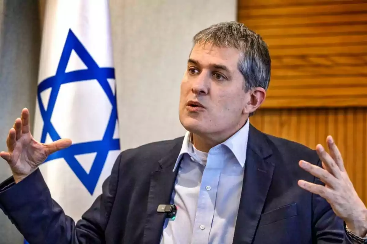 Gray-haired man in a blue jacket speaking and gesturing with his hands in front of an Israeli flag Gray-haired man in a blue jacket speaking and gesturing with his hands in front of an Israeli flag