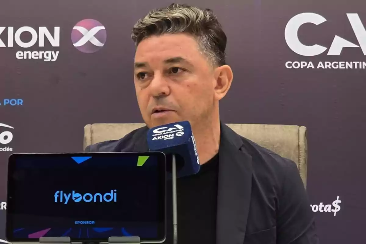 Short-haired, gray-haired man speaking at a press conference with a blue microphone and a screen displaying the Flybondi logo