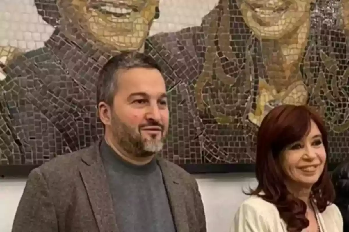 Dos personas posan sonrientes frente a un mural de mosaico que muestra rostros sonrientes Dos personas posan sonrientes frente a un mural de mosaico que muestra rostros sonrientes