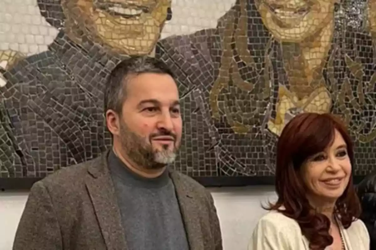 Dos personas posan frente a un mural de mosaico que muestra rostros sonrientes