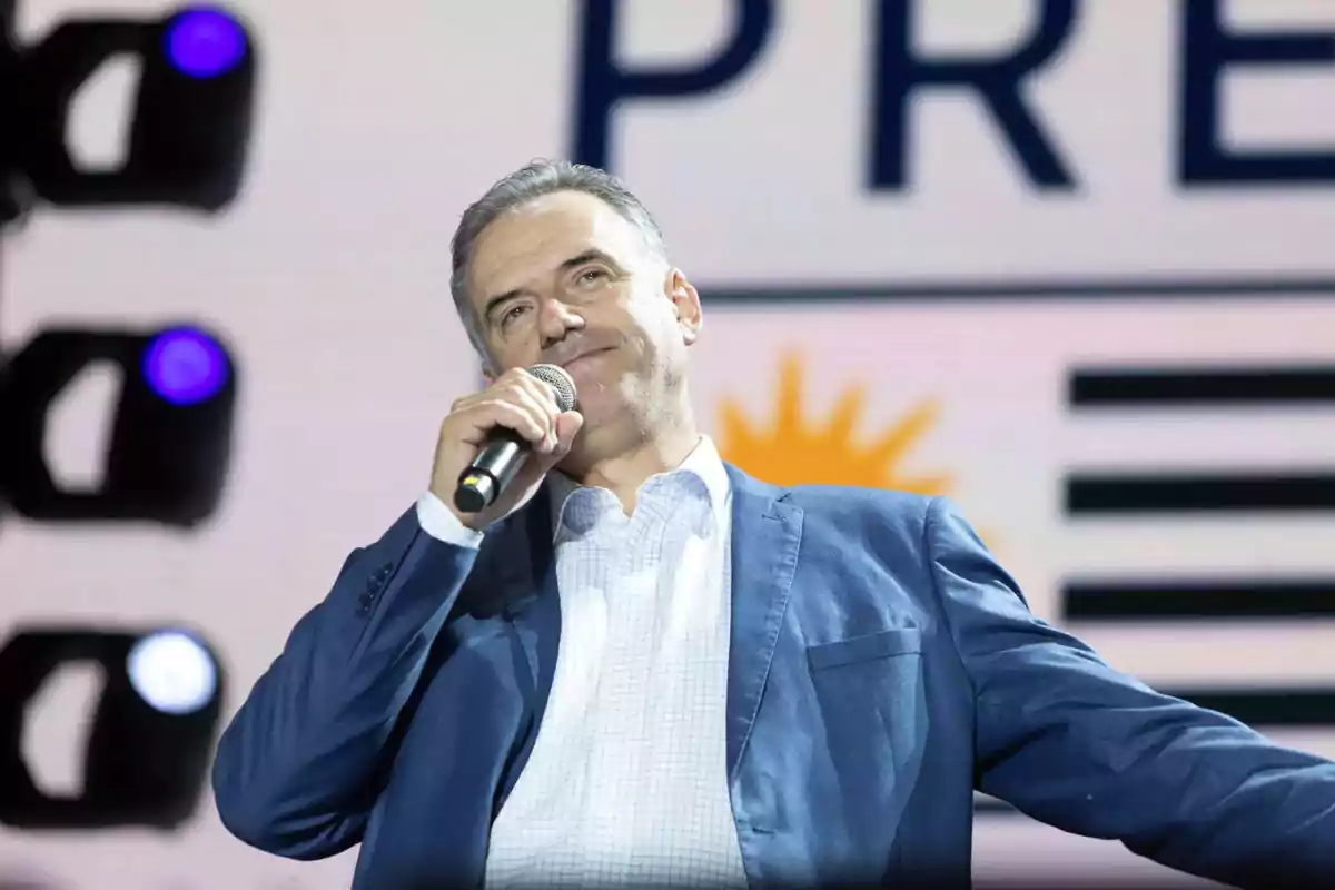 Hombre de traje azul hablando por micrófono en un escenario con fondo iluminado y un diseño de sol y franjas negras. Hombre de traje azul hablando por micrófono en un escenario con fondo iluminado y un diseño de sol y franjas negras.