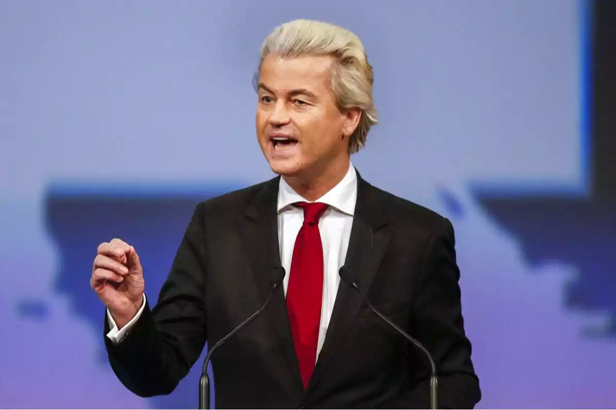 Blond-haired man in a dark suit speaking at a podium with a blurred blue background