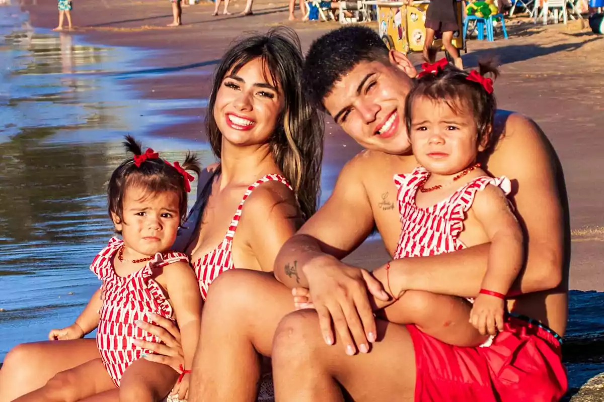 Smiling family sitting on the beach with two little girls wearing matching swimsuits and the sand and sea in the background