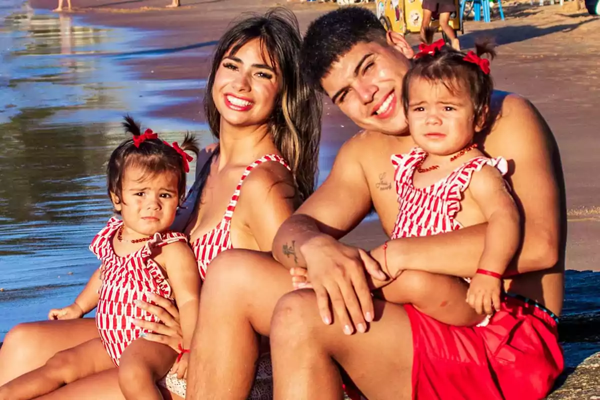 Familia sonriente sentada en la playa con dos niñas pequeñas usando trajes de baño a juego y la arena y el mar de fondo Familia sonriente sentada en la playa con dos niñas pequeñas usando trajes de baño a juego y la arena y el mar de fondo