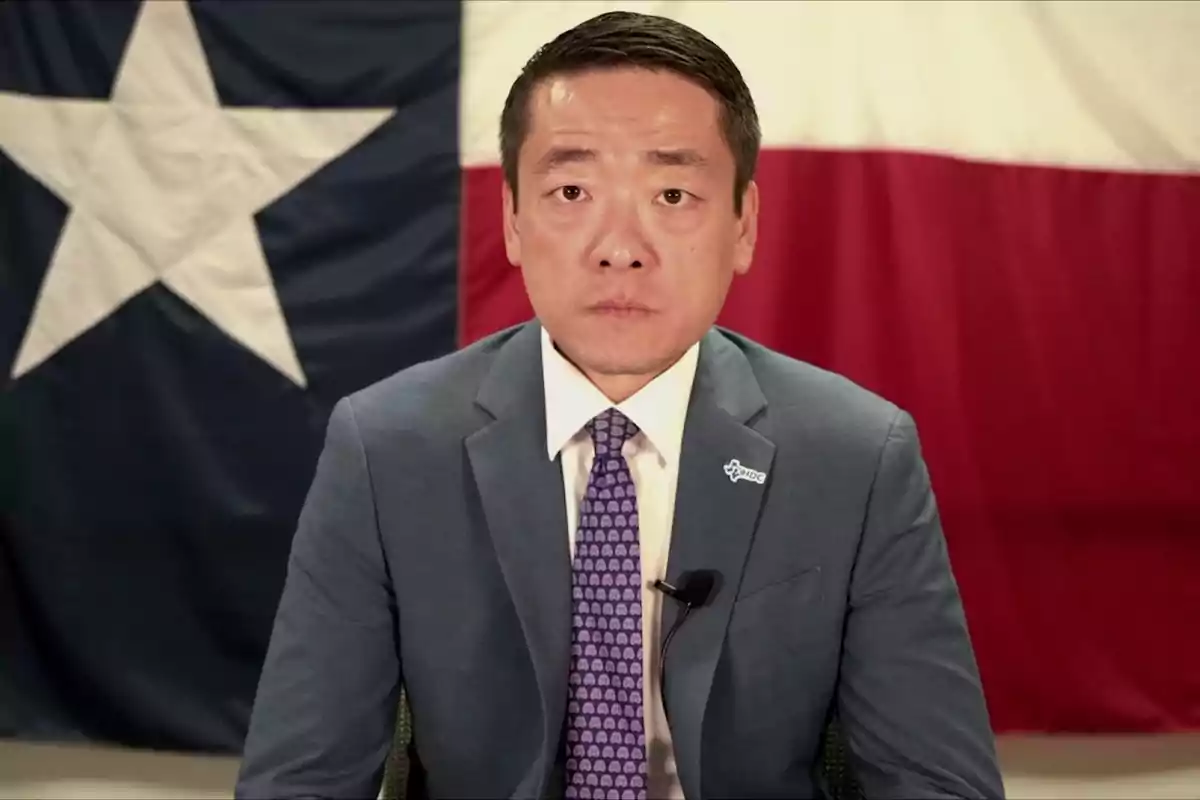 Hombre con traje azul y corbata morada sentado frente a una bandera de Texas