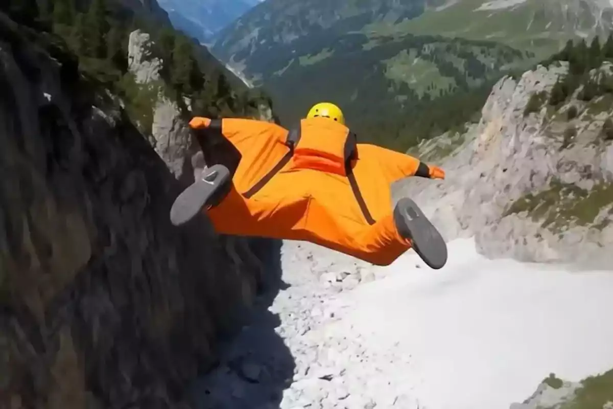 Persona con traje de alas naranja y casco amarillo volando entre montañas rocosas y verdes