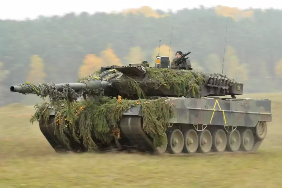 Tanque militar camuflado con ramas y follaje avanzando por un campo con un soldado visible en la torreta Tanque militar camuflado con ramas y follaje avanzando por un campo con un soldado visible en la torreta