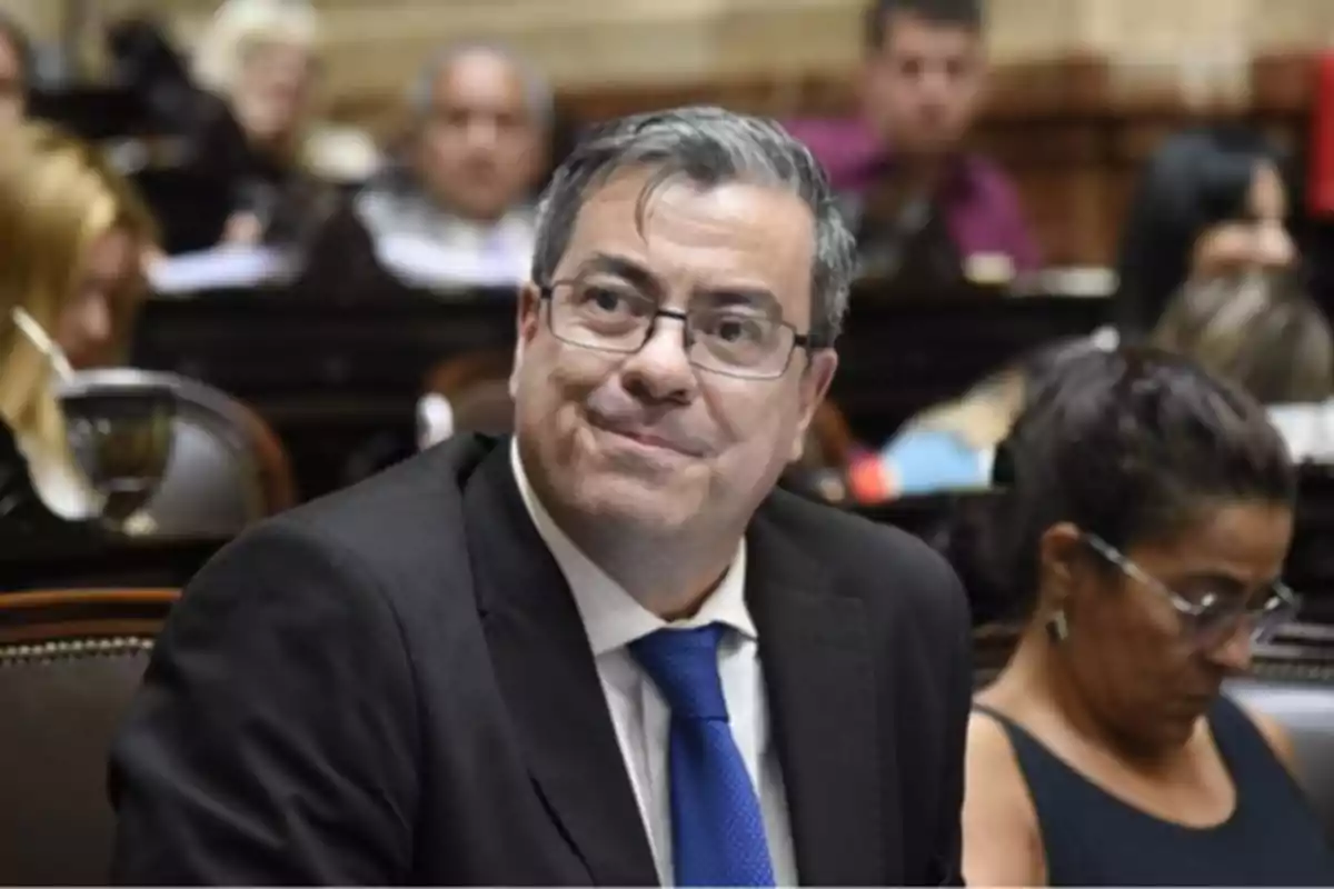 Hombre de cabello canoso y gafas con traje y corbata azul sentado en un recinto parlamentario rodeado de otras personas
