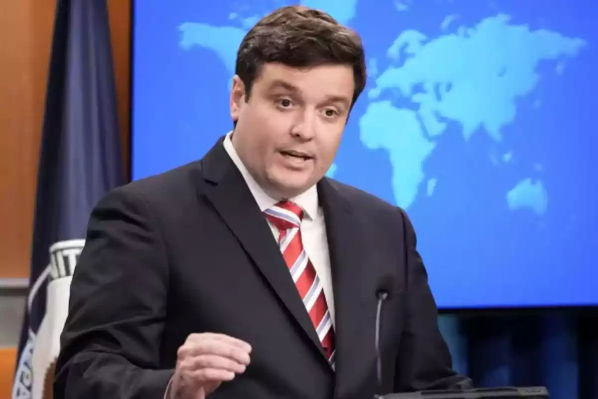 Short dark-haired man in a suit and tie speaking in front of a microphone with a blue background displaying a world map Short dark-haired man in a suit and tie speaking in front of a microphone with a blue background displaying a world map