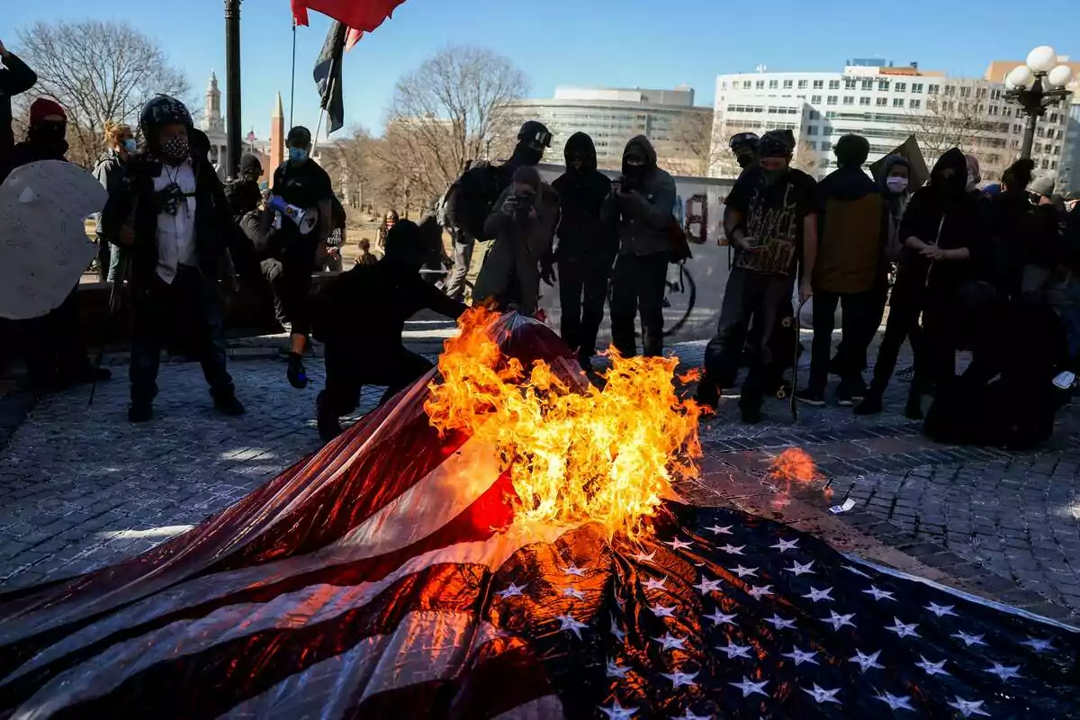 A group of people watches as a United States flag burns on the ground during a protest in an urban public space. A group of people watches as a United States flag burns on the ground during a protest in an urban public space.