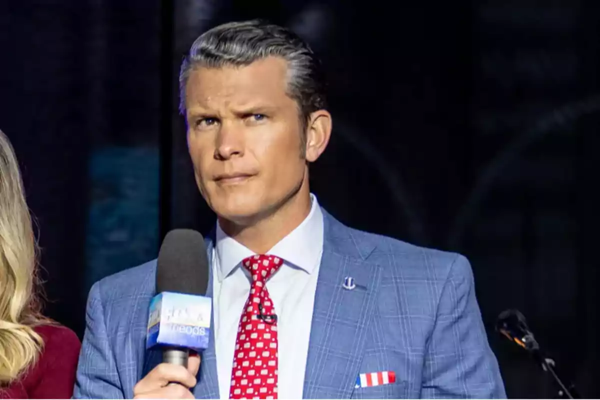 Gray-haired man with slicked-back hair, dressed in a blue suit and red tie, holding a Fox & Friends microphone and looking with a serious expression Gray-haired man with slicked-back hair, dressed in a blue suit and red tie, holding a Fox & Friends microphone and looking with a serious expression
