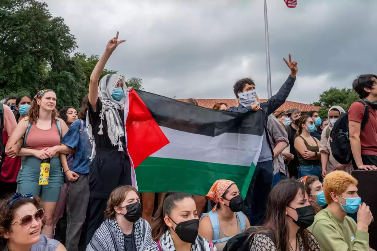 Grupo de personas manifestándose al aire libre, algunas con mascarillas y pañuelos, sosteniendo una bandera de Palestina y haciendo señales de paz con las manos