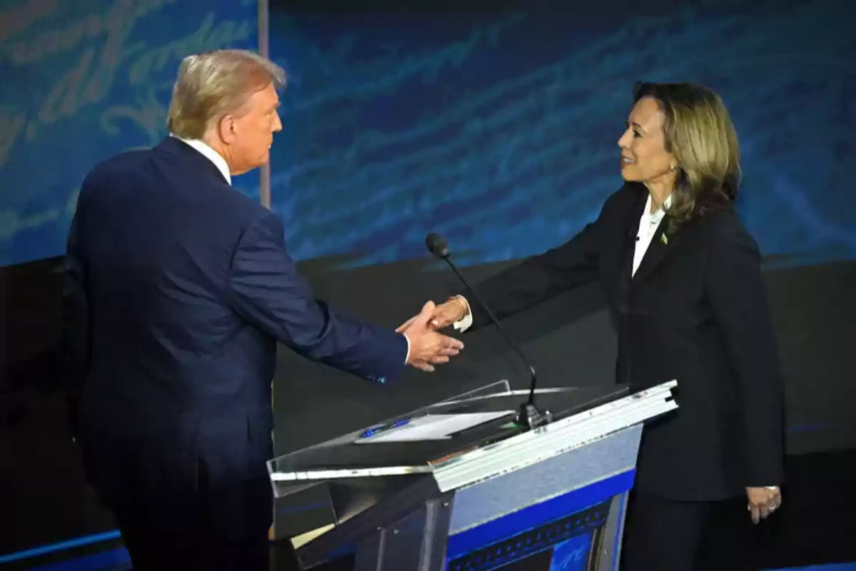 Two people in suits shake hands on a debate stage in front of a podium.