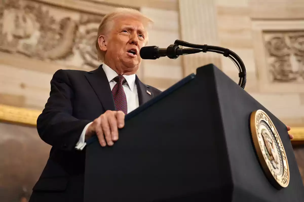 Blond-haired man in a dark suit and red tie speaking in front of a podium with the presidential seal of the United States in an elegant indoor setting