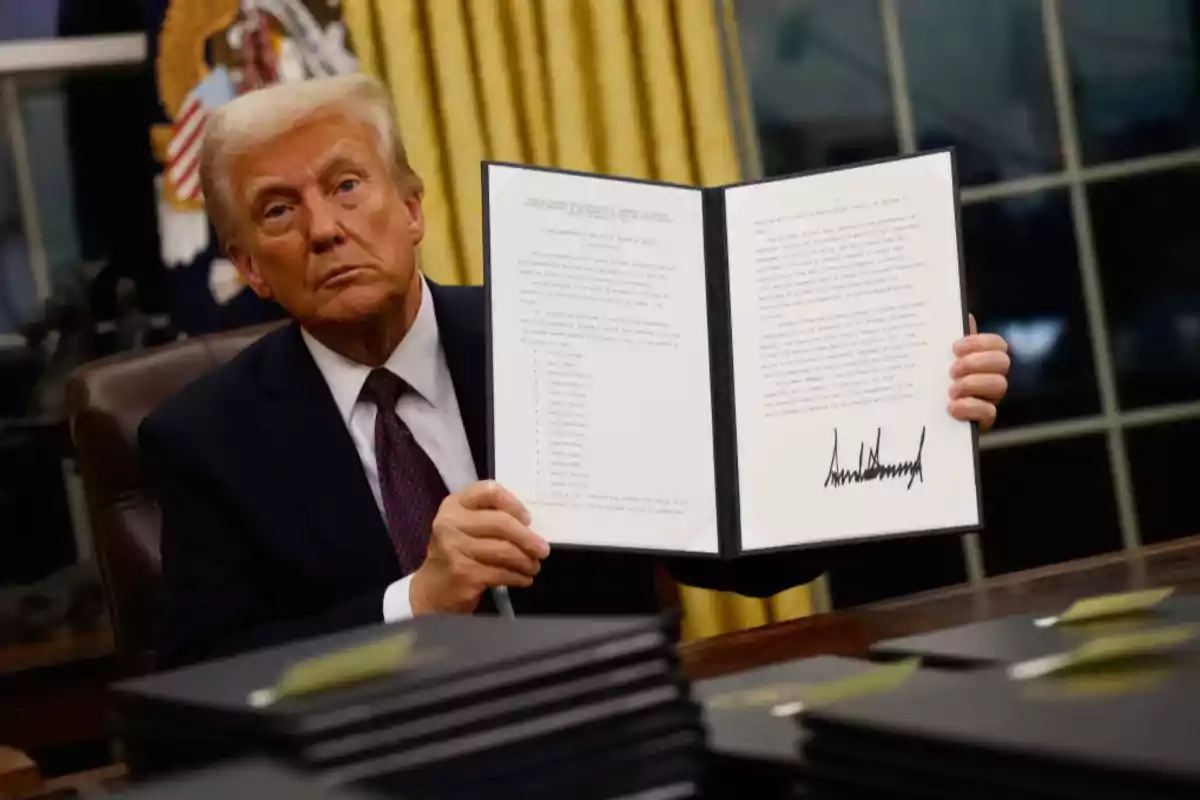 Blond-haired man in a dark suit and red tie showing a signed document in an office with golden curtains and the seal of the United States in the background Blond-haired man in a dark suit and red tie showing a signed document in an office with golden curtains and the seal of the United States in the background