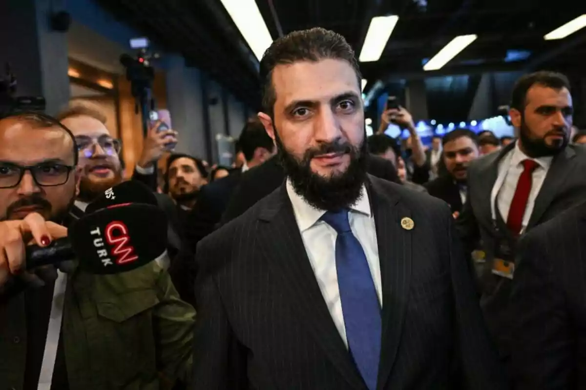 A man with a beard and a dark suit walks among a group of people and reporters while a CNN Turk camera and microphone focus on him at a crowded event. A man with a beard and a dark suit walks among a group of people and reporters while a CNN Turk camera and microphone focus on him at a crowded event.