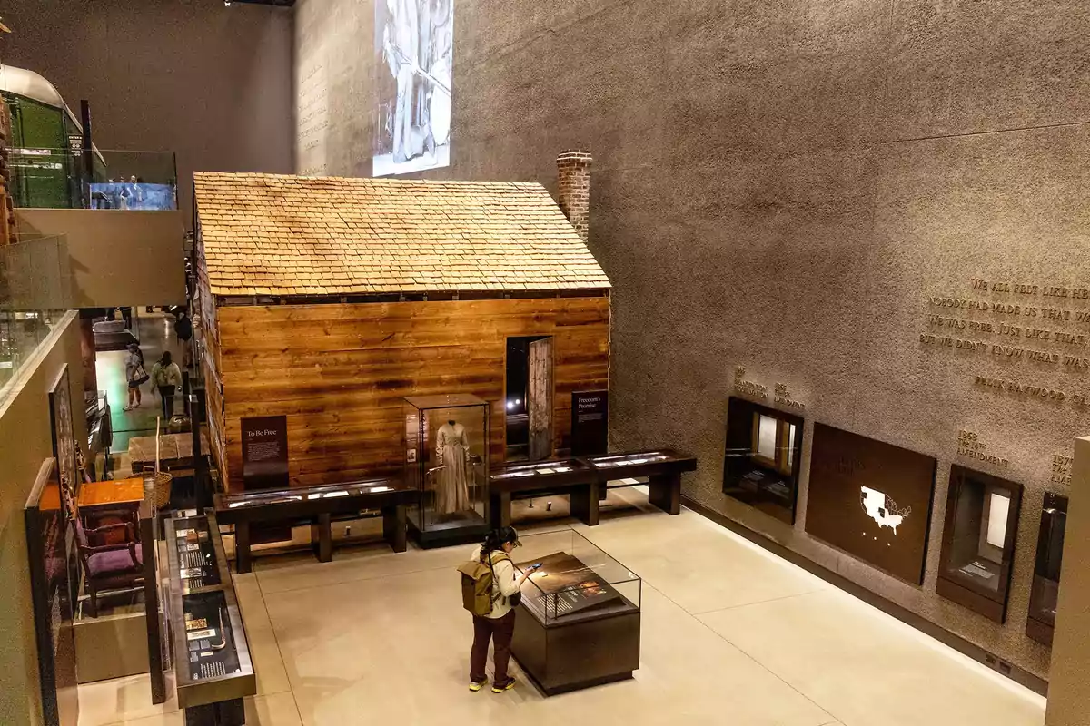 A person observes an exhibit in a museum where there is a replica of a wooden cabin surrounded by display cases with historical objects and informational panels on the walls. A person observes an exhibit in a museum where there is a replica of a wooden cabin surrounded by display cases with historical objects and informational panels on the walls.