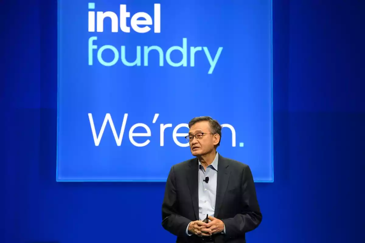 Hombre de traje hablando en un escenario con un fondo azul que muestra el logo de Intel Foundry y el texto We're in