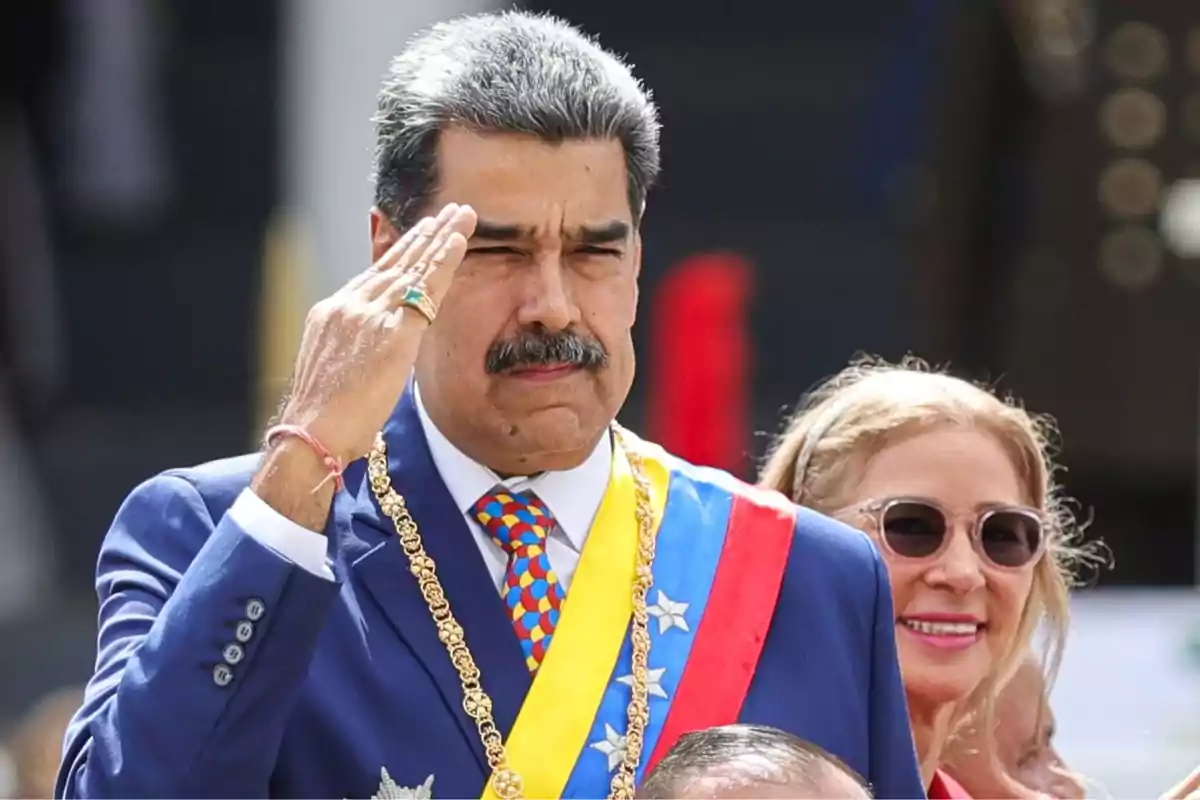 Man with a mustache and graying hair dressed in a blue suit and presidential sash giving a military salute while a woman wearing sunglasses smiles behind him Man with a mustache and graying hair dressed in a blue suit and presidential sash giving a military salute while a woman wearing sunglasses smiles behind him