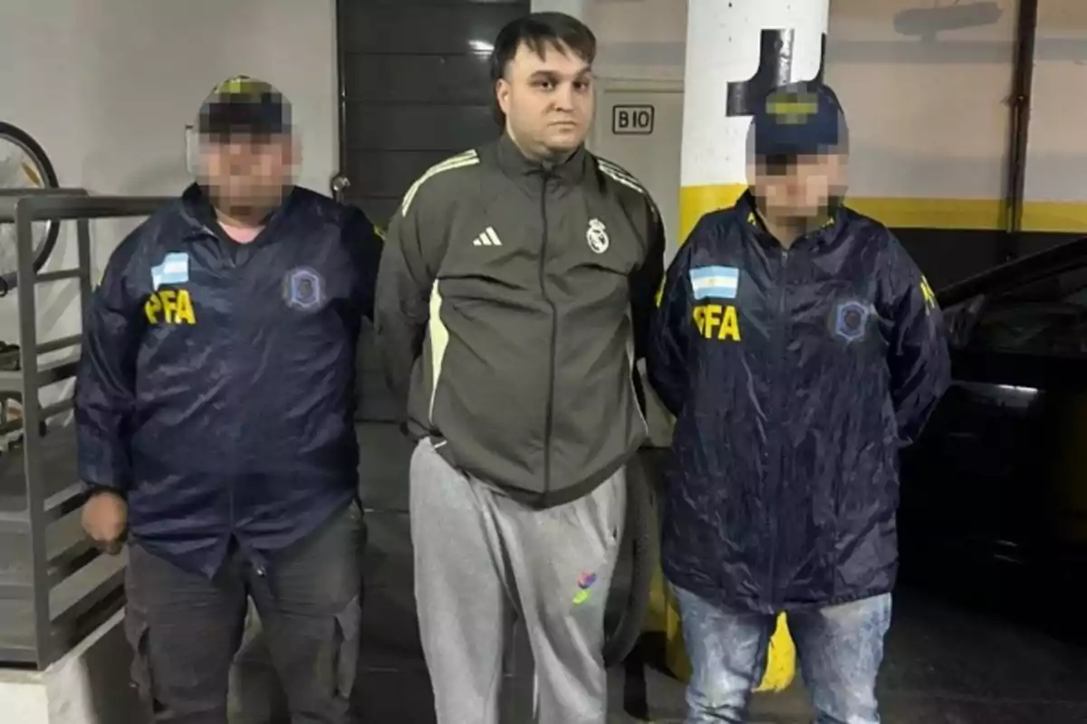 Tres personas de pie en un estacionamiento subterráneo, dos de ellas con chaquetas oscuras y gorras con las letras PFA y la persona del centro con ropa deportiva clara