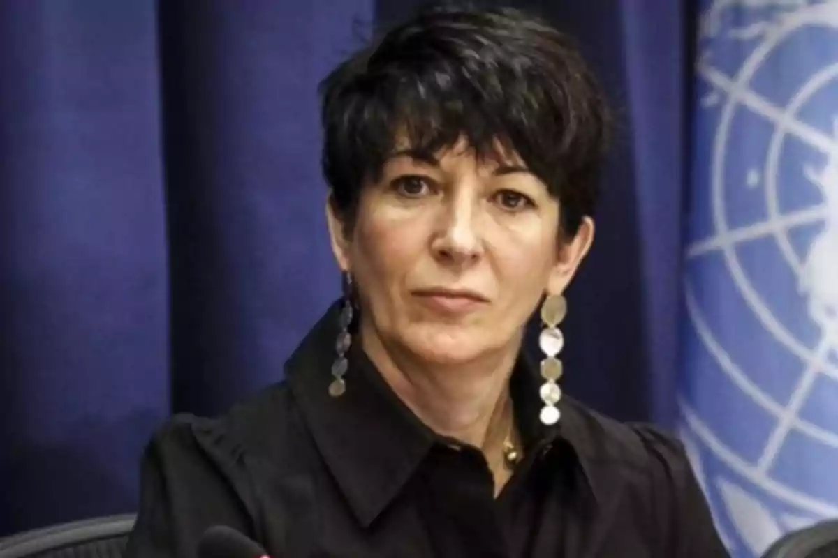 Short-haired, dark-haired woman with long earrings sitting in front of a blue background with an institutional logo