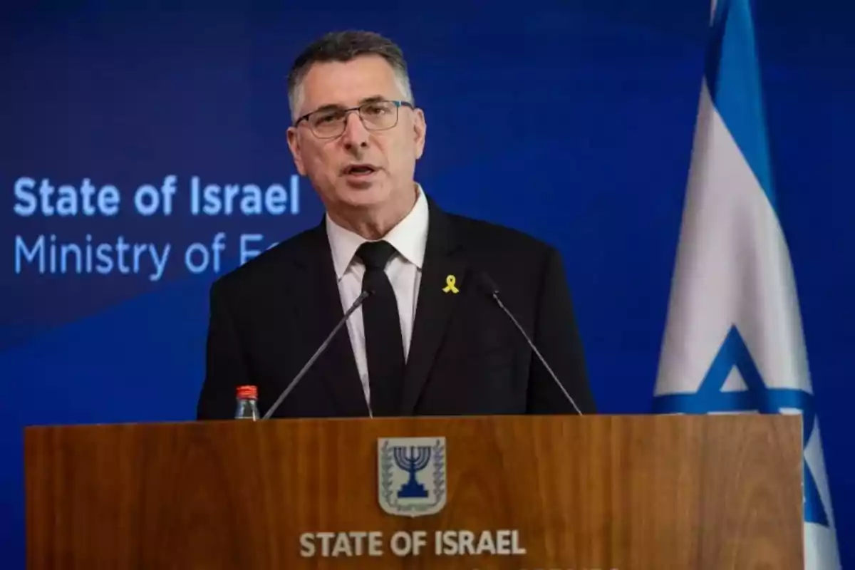 Hombre de traje oscuro y gafas hablando en un podio con el escudo de Israel y una bandera israelí al fondo