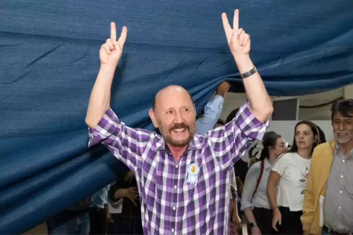 Hombre sonriente con camisa a cuadros morada levantando ambos brazos y haciendo el signo de la victoria con las manos mientras varias personas lo observan al fondo