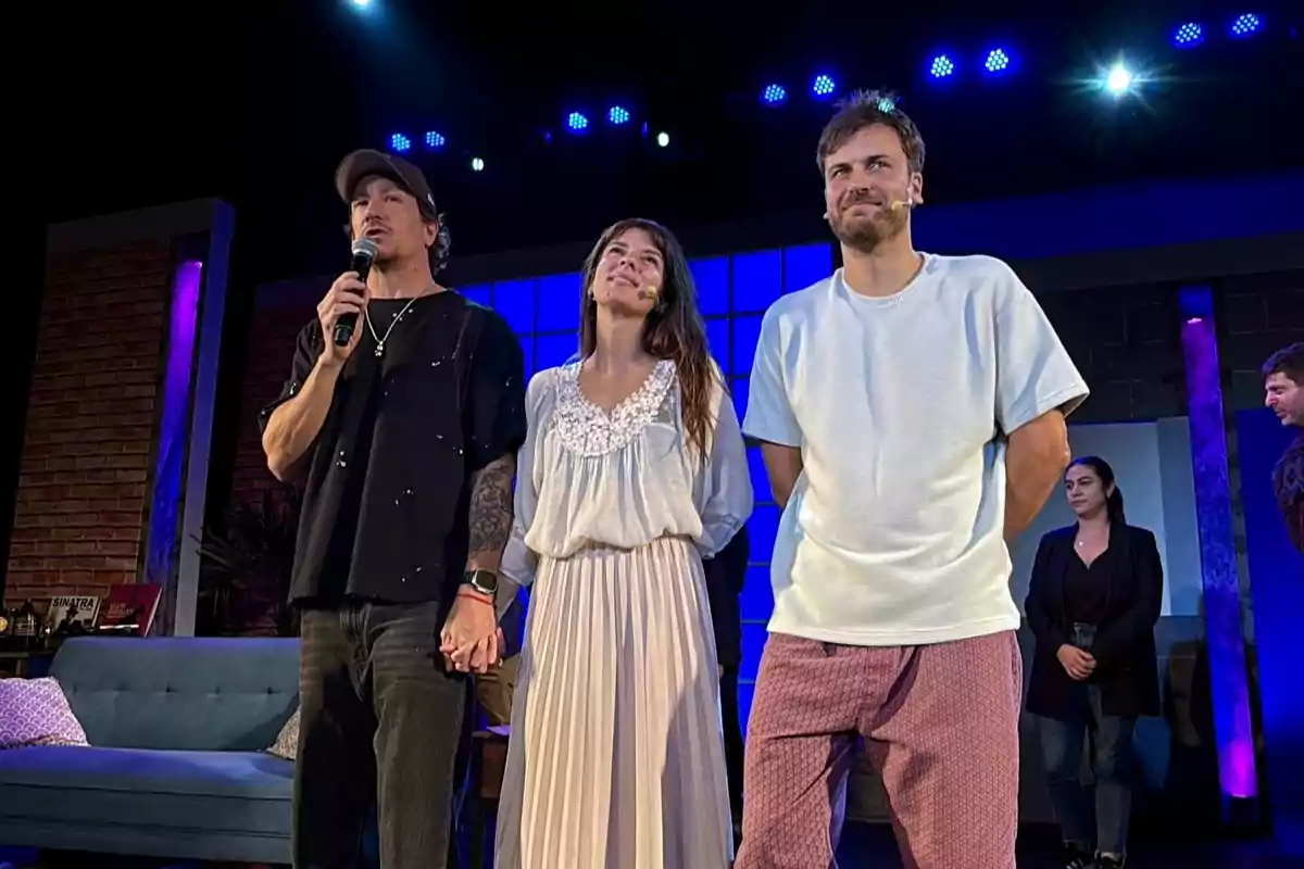 Three people are standing on a lit stage, one of them holding a microphone and another person's hand, while in the background two more people and a blue sofa can be seen. Three people are standing on a lit stage, one of them holding a microphone and another person's hand, while in the background two more people and a blue sofa can be seen.