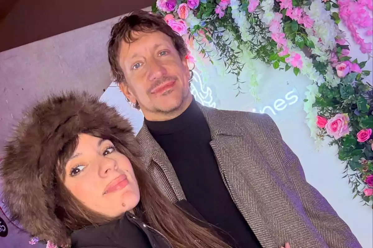 Una mujer con gorro de peluche y un hombre con saco posan juntos frente a una pared decorada con flores rosas y blancas.