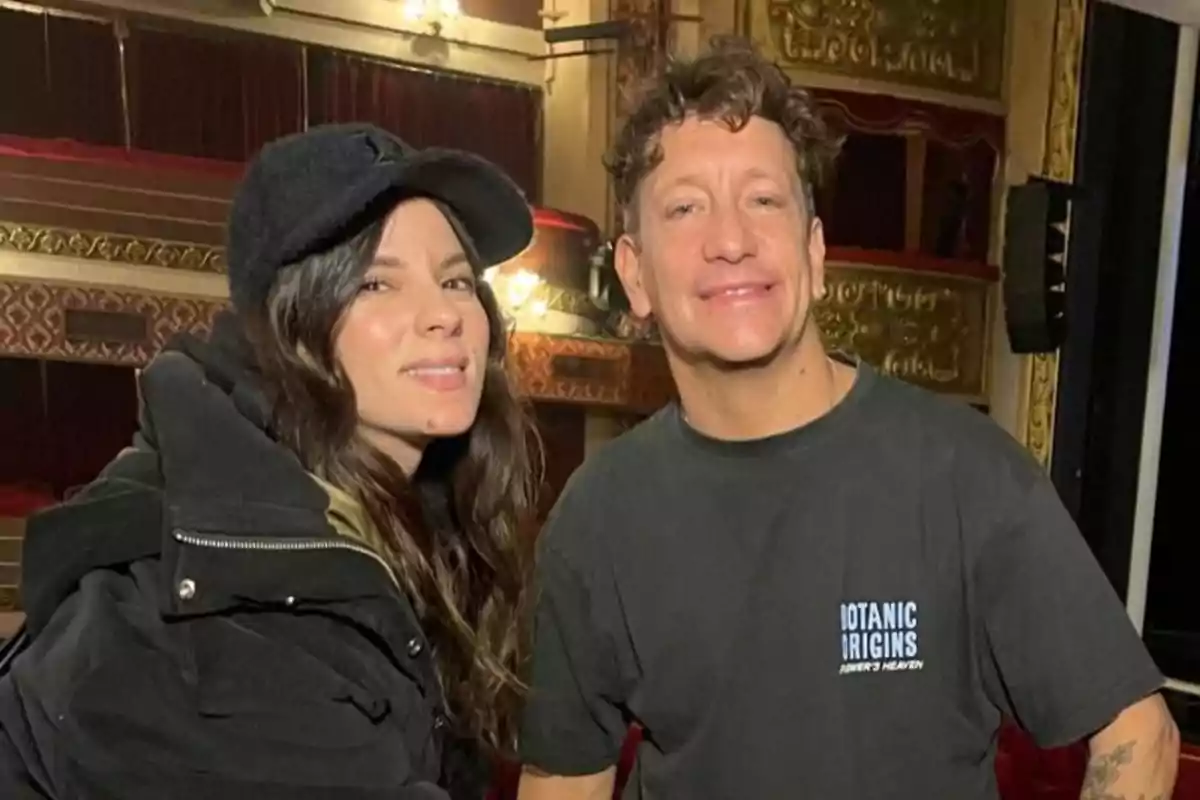 Two people pose smiling inside a theater with golden decorations and red curtains in the background. Two people pose smiling inside a theater with golden decorations and red curtains in the background.