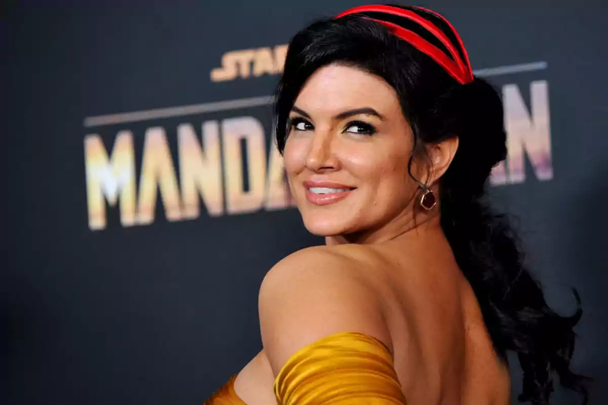 Mujer con vestido dorado y diadema roja sonriendo en una alfombra roja frente a un cartel de The Mandalorian Mujer con vestido dorado y diadema roja sonriendo en una alfombra roja frente a un cartel de The Mandalorian