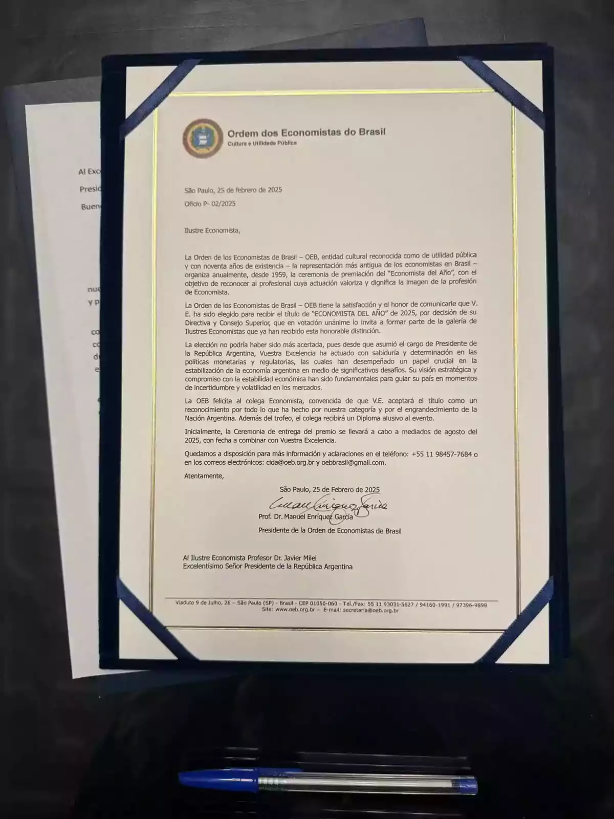 Carta oficial de la Orden de los Economistas de Brasil dirigida a Javier Milei, anunciando su elección como Economista del Año 2025, firmada por el presidente de la organización y presentada en un portadiplomas sobre una mesa junto a una pluma y otro documento parcialmente visible.