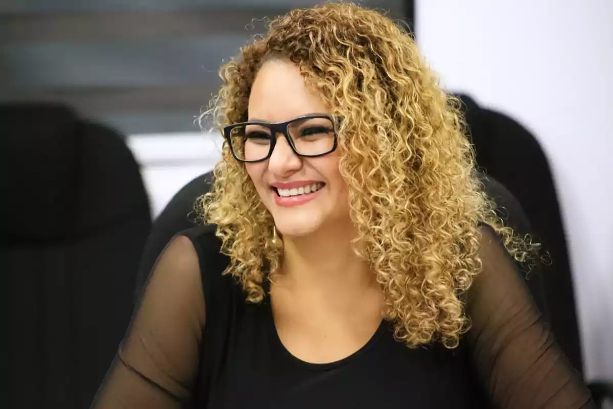 Mujer de cabello rizado y rubio usando gafas negras y blusa negra sonríe mientras está sentada en una oficina Mujer de cabello rizado y rubio usando gafas negras y blusa negra sonríe mientras está sentada en una oficina