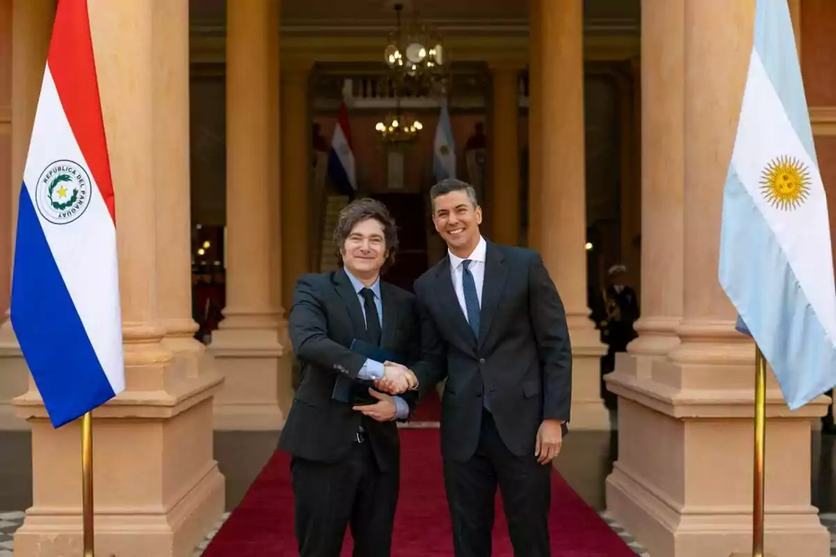 Two men in suits shake hands and smile in front of an official building, with the flags of Paraguay and Argentina on either side. Two men in suits shake hands and smile in front of an official building, with the flags of Paraguay and Argentina on either side.