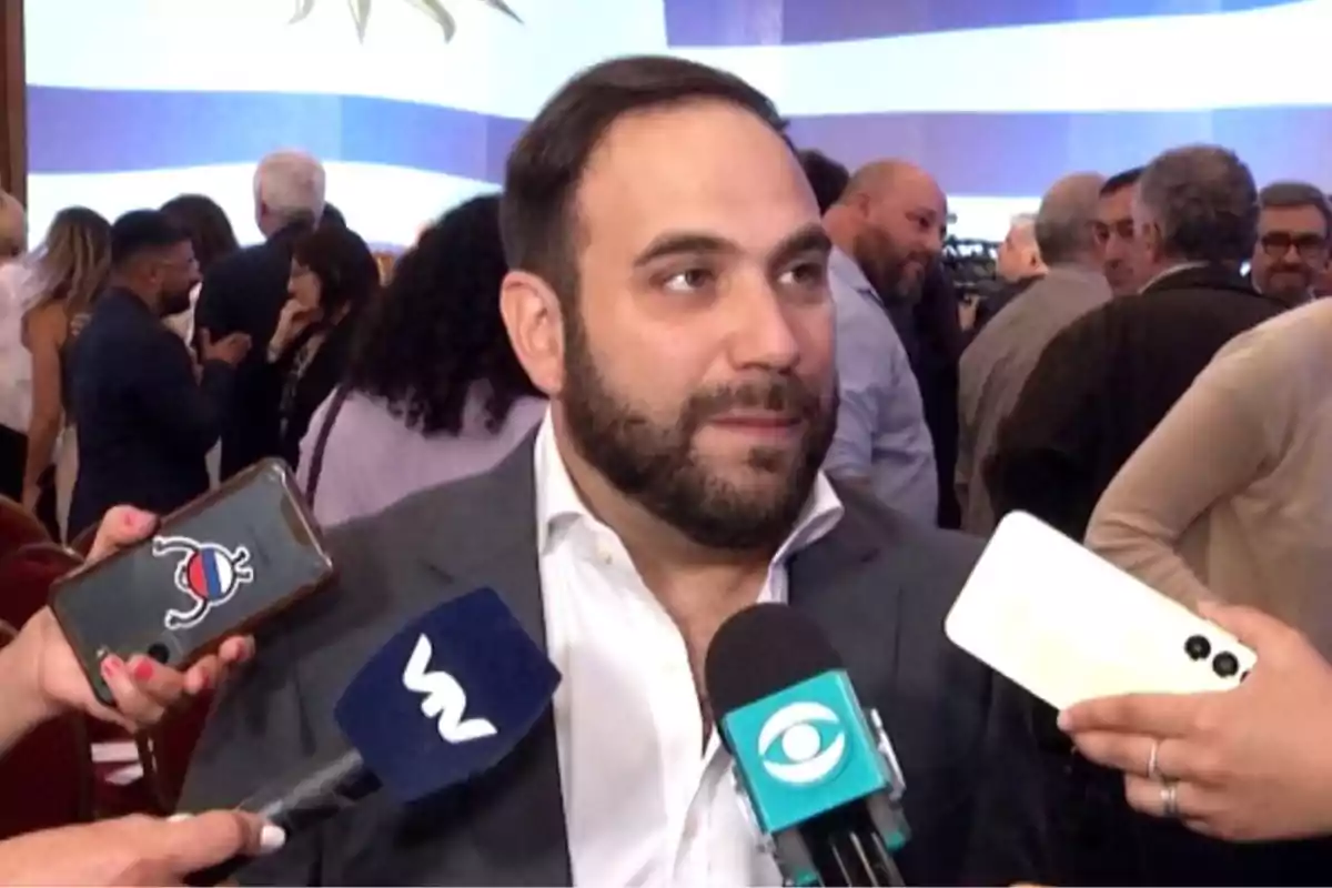 A bearded man in a suit is interviewed by several media outlets at a crowded event. A bearded man in a suit is interviewed by several media outlets at a crowded event.