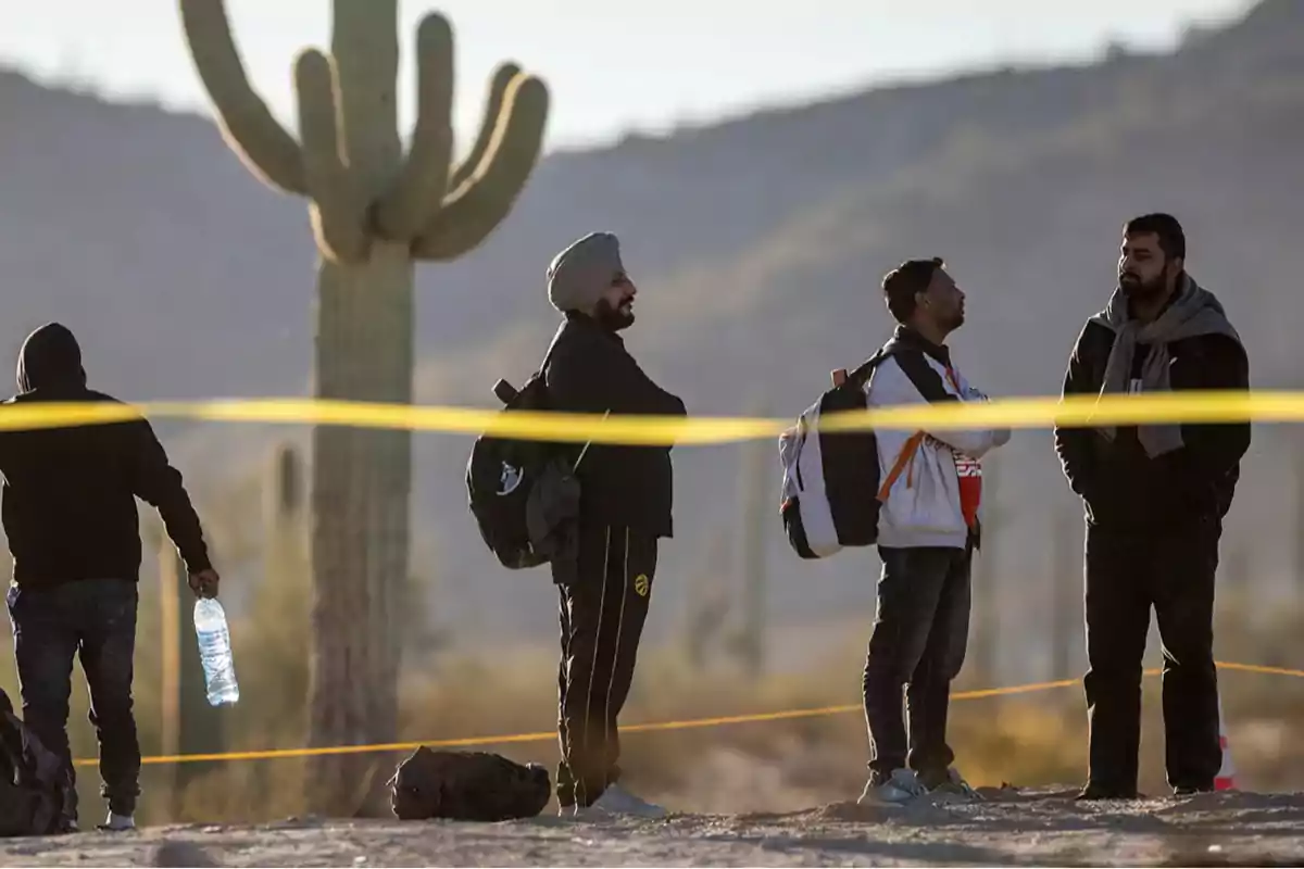 A group of people with backpacks waits in a desert area next to a large cactus and a yellow tape, with mountains in the background. A group of people with backpacks waits in a desert area next to a large cactus and a yellow tape, with mountains in the background.