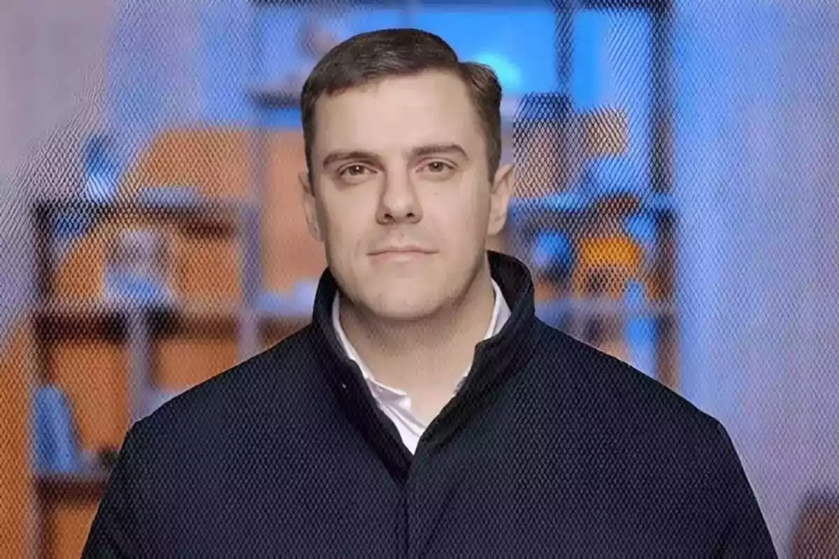 Hombre de cabello corto y oscuro con chaqueta negra posando frente a un fondo desenfocado de tonos azules y naranjas Hombre de cabello corto y oscuro con chaqueta negra posando frente a un fondo desenfocado de tonos azules y naranjas
