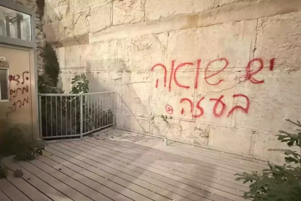 Pared de piedra antigua con grafiti en hebreo pintado con aerosol rojo y una barandilla metálica junto a una pequeña planta