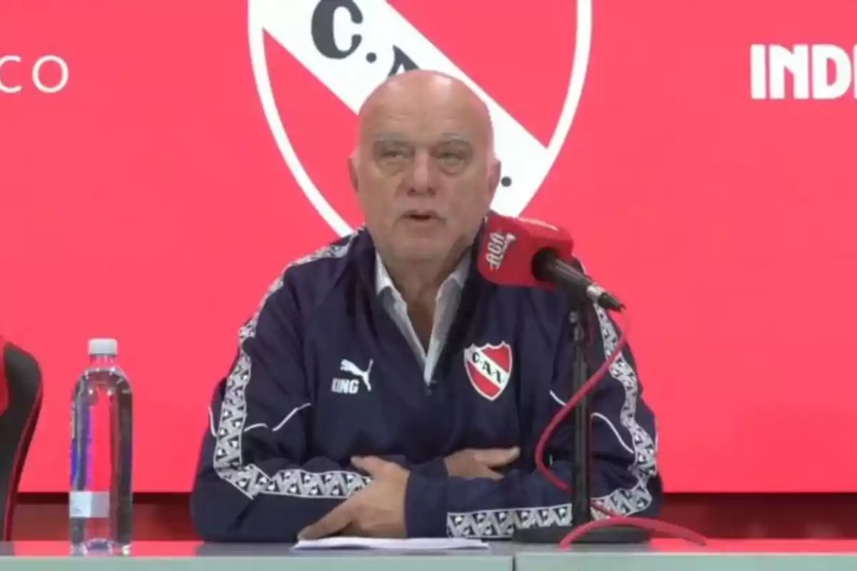 Older man wearing an Independiente sports jacket speaking at a press conference in front of a red microphone and a red background with the club’s crest.