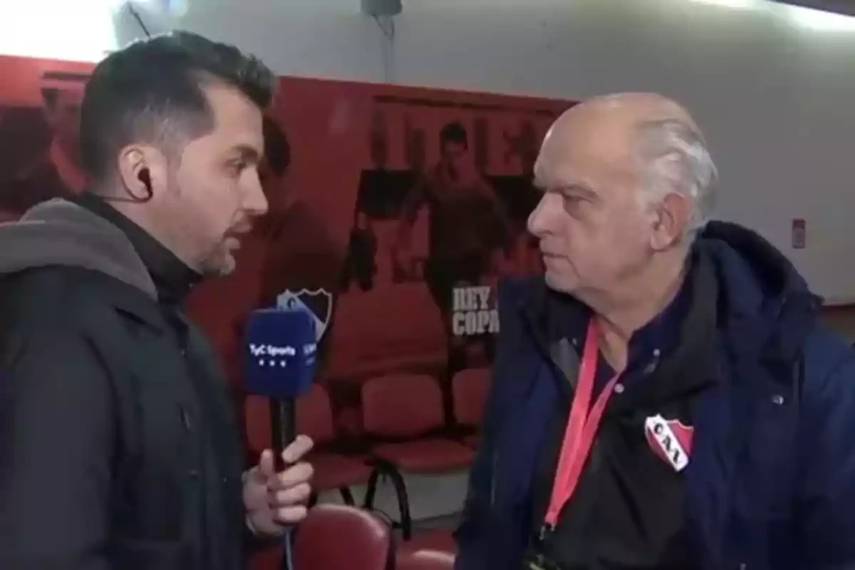 A journalist interviews an older man who is wearing a badge and a jacket with a soccer club crest in an indoor setting. A journalist interviews an older man who is wearing a badge and a jacket with a soccer club crest in an indoor setting.