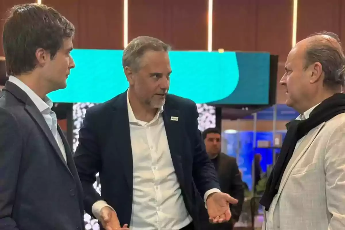 Three men dressed in suits are talking at a business event in an indoor setting with screens and lights in the background. Three men dressed in suits are talking at a business event in an indoor setting with screens and lights in the background.