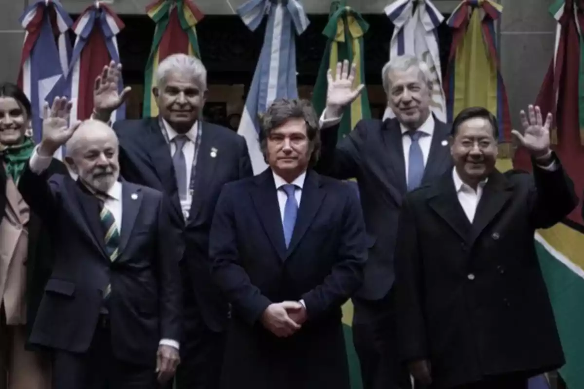 Group of political leaders posing together in front of flags from various countries while some of them wave with their hands raised