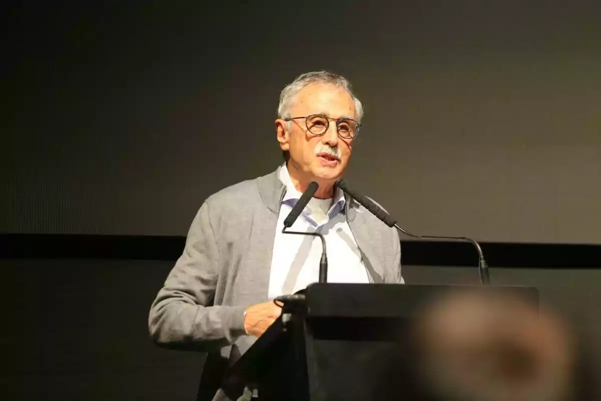 Older man with glasses and gray hair speaking at a podium with two microphones in an indoor setting