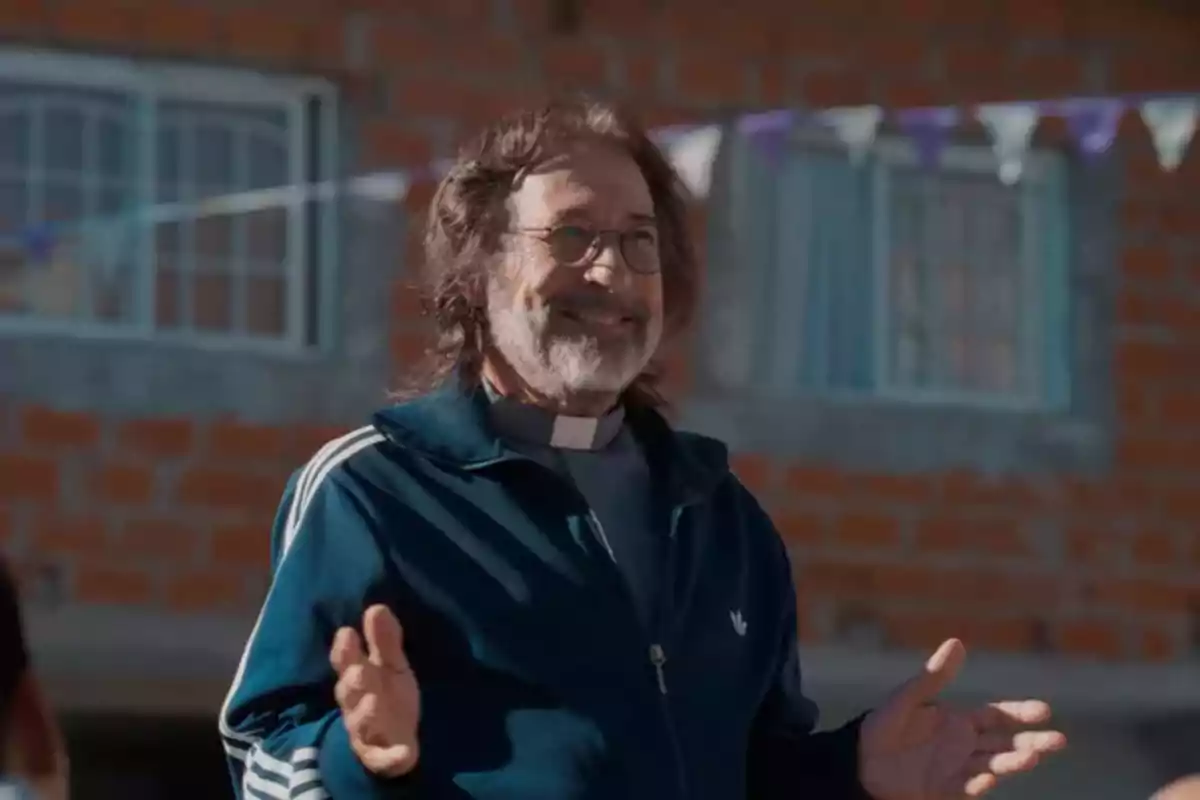 Older man with glasses and a beard smiles while wearing a blue tracksuit and a clerical collar, standing in front of a brick house with windows and pennants in the background. Older man with glasses and a beard smiles while wearing a blue tracksuit and a clerical collar, standing in front of a brick house with windows and pennants in the background.
