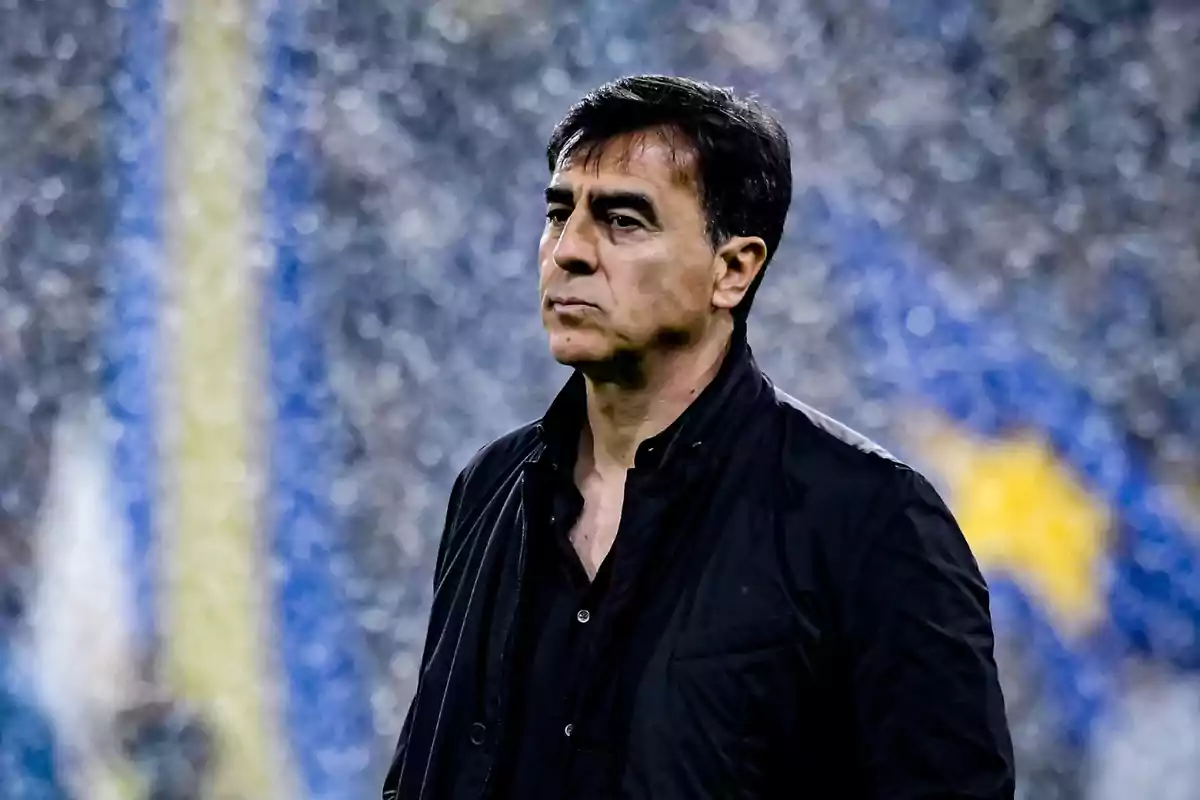 Hombre de cabello oscuro con expresión seria vistiendo chaqueta negra en un entorno deportivo desenfocado Hombre de cabello oscuro con expresión seria vistiendo chaqueta negra en un entorno deportivo desenfocado