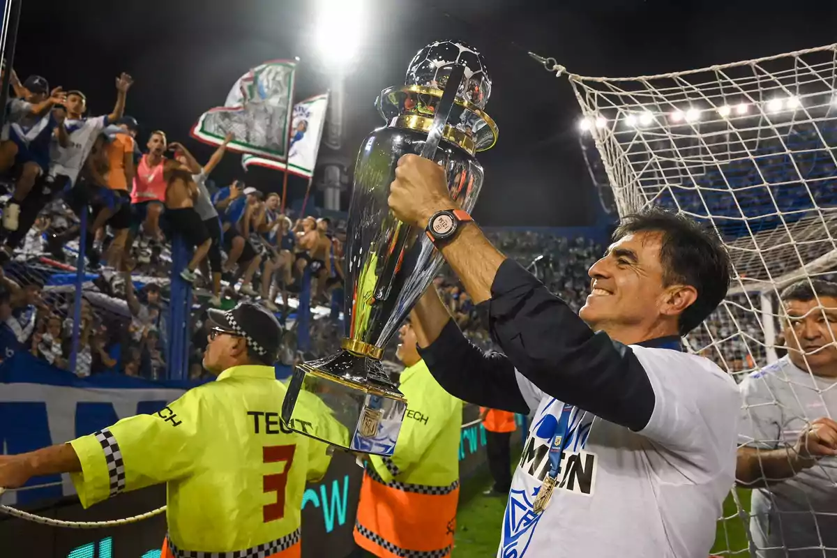 Hombre sonriente levantando un trofeo de fútbol mientras celebra con aficionados y personal de seguridad en un estadio lleno Hombre sonriente levantando un trofeo de fútbol mientras celebra con aficionados y personal de seguridad en un estadio lleno