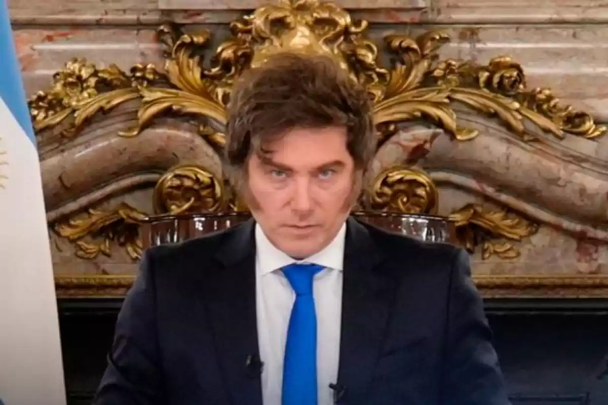 Hombre de cabello castaño con traje oscuro y corbata azul sentado frente a un fondo decorativo dorado y mármol