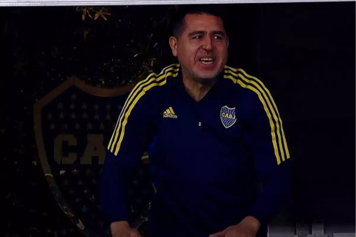 Hombre con chaqueta azul y amarilla de Boca Juniors sonriendo frente a un escudo del club